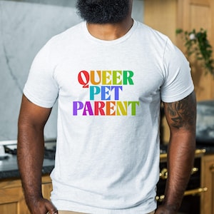 Puede incluir: Camiseta gris claro con el texto "QUEER PET PARENT" en colores arcoíris. La camiseta es de cuello redondo y parece estar hecha de un material suave. El texto está centrado en la parte delantera de la camiseta.