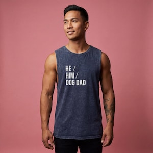 May include: A navy blue sleeveless shirt with the text "HE / HIM / DOG DAD" in white. The shirt has a faded, textured appearance. The background is a solid pink color.