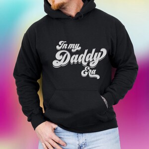 Pode incluir: Camisola de moletom com capuz preta com o texto "In my Daddy Era" em letras brancas de estilo retrô.