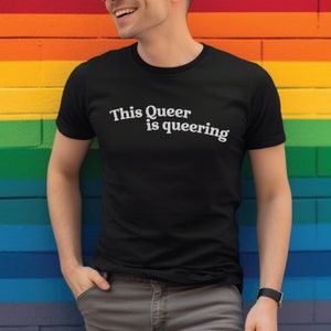 Puede incluir: Una persona lleva una camiseta negra con el texto "This Queer is queering" en letras blancas. La persona está de pie frente a una pared de ladrillo de colores arcoíris.