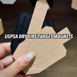 Può includere: Un set di magneti per bersagli da tiro a secco USPSA in nero e beige. Il bersaglio beige presenta zone di punteggio in rilievo contrassegnate con A, C e D, con un rettangolo per la zona A. I bersagli neri sono parzialmente visibili dietro. Il testo USPSA DRY FIRE TARGET MAGNETS è sovrapposto.