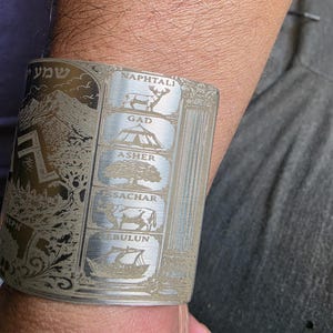 May include: A silver-toned metal cuff bracelet featuring intricate laser-etched designs. The surface displays vertical panels with the names Naphtali, Gad, Asher, Issachar, and Zebulun, each accompanied by a corresponding icon of a deer, tent, tree, ox, and ship.