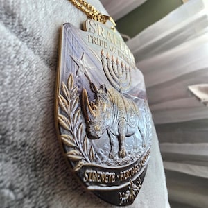 May include: A gold-colored pendant with a rhino design, the Star of David, and a menorah. The pendant is on a gold chain. The text on the pendant reads "ISRAELI TRIBE OF EPHRAIM" and "STRENGTH HERITAGE COMMUNITY".