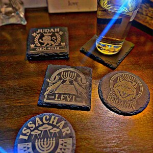 May include: A collection of engraved slate coasters with religious symbols and text. The coasters feature designs for Judah, Levi, Issachar, and Benjamin. A glass of liquid sits on one of the coasters. The coasters are on a wooden surface.