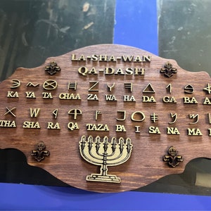 May include: Wooden plaque with the words "LA-SHA-WAN QA-DASH" and various symbols and letters. The plaque features a menorah and fleur-de-lis designs. The background is a dark wood color.