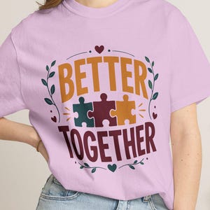 Könnte beinhalten: Ein hellviolettes Kurzarm-T-Shirt mit einem Grafikdesign und dem Text BETTER TOGETHER in Orange und Dunkelrot. Das Design zeigt drei ineinandergreifende Puzzleteile in Petrol, Rot und Gelb, umgeben von kleinen Blatt- und Herzmotiven.