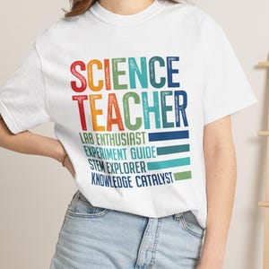 Könnte beinhalten: Ein weißes kurzärmeliges T-Shirt mit dem Text SCIENCE TEACHER in einer mehrfarbigen Verlaufsschrift, gefolgt von den Phrasen LAB ENTHUSIAST, EXPERIMENT GUIDE, STEM EXPLORER und KNOWLEDGE CATALYST in verschiedenen Blau- und Grüntönen.