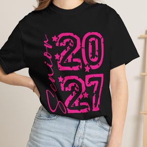 Könnte beinhalten: Ein schwarzes Kurzarm-T-Shirt mit dem vertikal in pinker Schreibschrift gedruckten Wort Senior sowie den großen, pinken, abgenutzt wirkenden Zahlen 2027, die mit kleinen Sternen verziert sind. Das Design ist mittig auf der Vorderseite platziert.