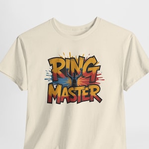Könnte beinhalten: Ein cremefarbenes Kurzarm-T-Shirt mit einem Grafikdruck, der den Text RING MASTER in fetten gelben und roten Buchstaben zeigt. Das Design enthält die Silhouette einer Figur mit erhobenen Armen vor einem bunten blauen und roten Farbspritzer.