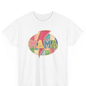 Könnte beinhalten: Ein weißes T-Shirt mit kurzen Ärmeln und einem zentralen Aufdruck des Wortes MAMA in Blockbuchstaben. Der Schriftzug ist mit einem pinken Blitz überlagert und befindet sich auf einem ovalen Hintergrund aus bunten Stoffmustern.