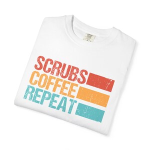 Könnte beinhalten: Ein weißes Kurzarm-T-Shirt mit einem abgenutzten Grafikdruck, der den Text "SCRUBS COFFEE REPEAT" in gestapelten Zeilen zeigt. Jedes Wort wird von einem horizontalen rechteckigen Block in Rot, Orange beziehungsweise Blaugrün begleitet.