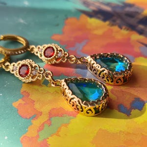 Puede incluir: Un par de pendientes de oro con piedras preciosas azules y verdes en forma de lágrima. Los pendientes tienen un delicado diseño de filigrana y están adornados con pequeñas piedras preciosas rojas.