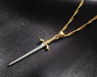 May include: A gold-colored necklace with a pendant shaped like a miniature sword. The sword pendant has a silver-colored blade and gold-colored hilt. The necklace chain is a twisted rope design, and the background is a dark, textured fabric.