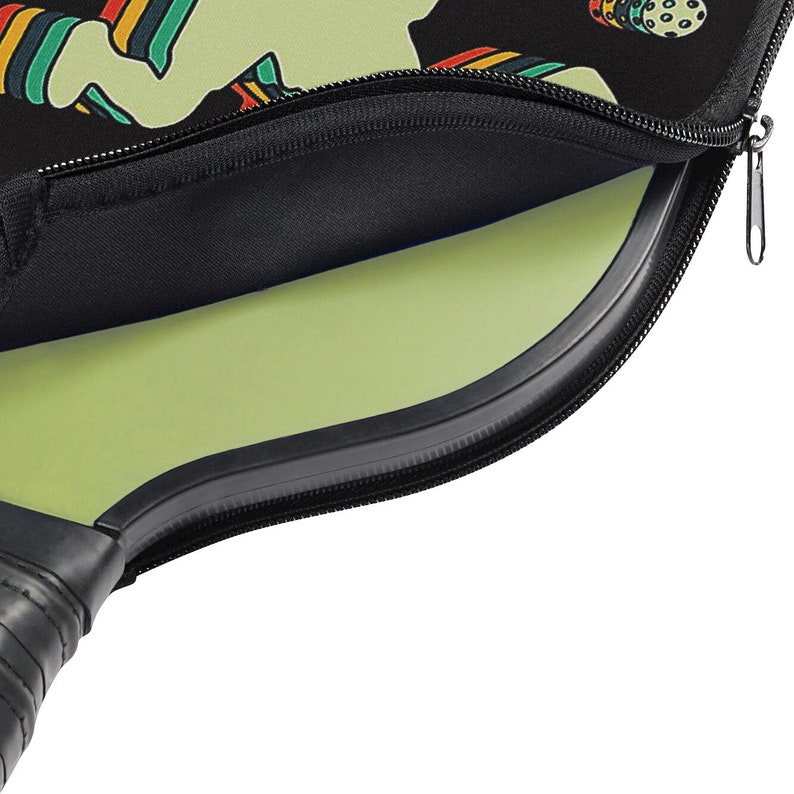 May include: A black zippered carrying case with a green paddle inside. The case has a colorful design with a person playing pickleball.