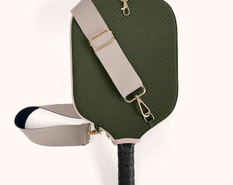 May include: A green pickleball paddle with a perforated surface and a tan shoulder strap. The paddle has a black grip and gold-colored hardware. The edges are trimmed with a light pink color.