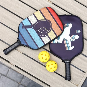 May include: Two colorful pickleball paddles with a retro striped design and a black paddle with a white silhouette of a player.  The paddles are on a wooden surface with two yellow pickleballs.