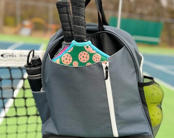 May include: A gray backpack with a large zippered pocket on the front. Two pickleball paddles are stored in the pocket. The backpack has a mesh side pocket with pickleballs inside.