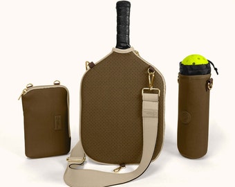 Könnte beinhalten: Eine braune Pickleball-Schlägerhülle, eine passende Zubehörtasche und ein Ballhalter. Die Schlägerhülle hat ein perforiertes Design und einen beigen Riemen. Der Ballhalter enthält einen gelben Pickleball. Alle Artikel haben goldfarbene Hardware.
