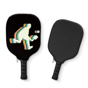 May include: Two black pickleball paddles. One paddle has a retro-style graphic of a person playing pickleball. The graphic is in shades of orange, yellow, green, and blue.