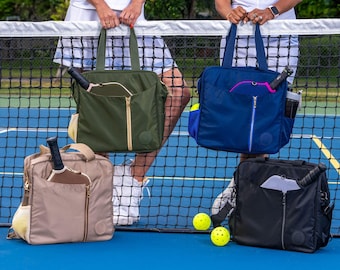 May include: Four different colored pickleball bags hanging on a tennis net. The bags are all made of a nylon material and have a zipper closure. Each bag has a pocket on the front and a handle on the top. The bags are hanging on a black tennis net with a blue court behind it.