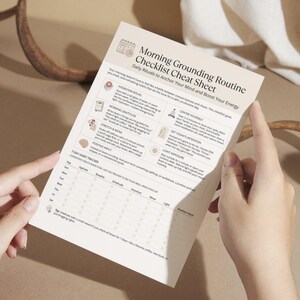Hands holding a printed copy of the Morning Grounding Routine Checklist cheat sheet with clear sections for rituals and a weekly tracker.