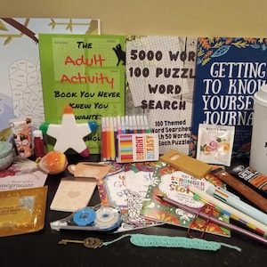 May include: A collection of items including books titled The Adult Activity Book, 5000 Word 100 Puzzle Word Search, and Getting To Know Yourself Journal, a white cup with Just One More Cup text, colored markers, a star-shaped object, and various stationery.