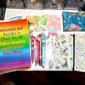May include: A collection of stationery items including a rainbow-colored book titled Activities and Puzzles to Cheer You UP Relieve Anxiety and Brighten Your Mood by Tamara L. Adams, several pens, a floral patterned pouch, and a sheet of daisy stickers.
