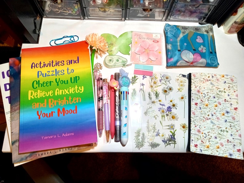 May include: A collection of stationery items including a rainbow-colored book titled Activities and Puzzles to Cheer You UP Relieve Anxiety and Brighten Your Mood by Tamara L. Adams, several pens, a floral patterned pouch, and a sheet of daisy stickers.