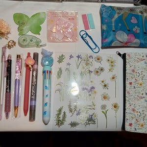 May include: A collection of stationery items including a flower-topped pen, a green butterfly-shaped sticky note, a cherry blossom sticker pack, a blue paperclip, a floral pouch, a multi-color pen, and a sheet of various botanical stickers.