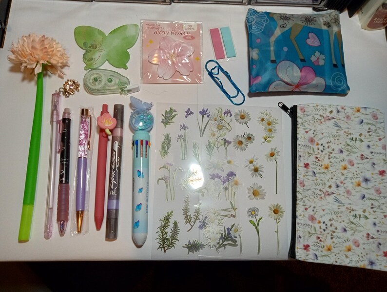 May include: A collection of stationery items including a flower-topped pen, a green butterfly-shaped sticky note, a cherry blossom sticker pack, a blue paperclip, a floral pouch, a multi-color pen, and a sheet of various botanical stickers.