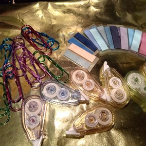 May include: A collection of stationery supplies including metallic heart-shaped paper clips in blue, purple, and green, a set of rectangular adhesive page markers in various pastel and dark shades, and several clear plastic correction tape dispensers.