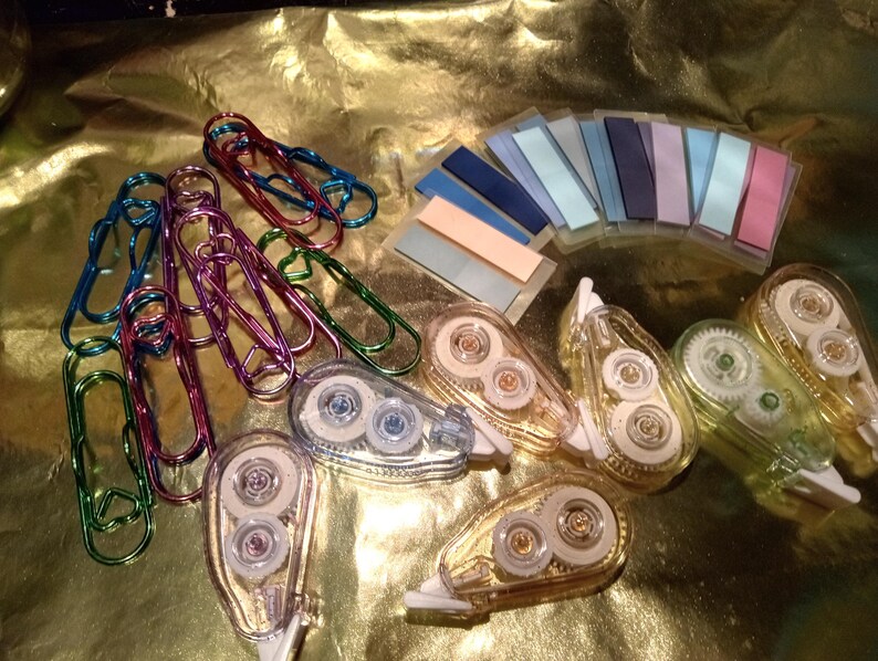 May include: A collection of stationery supplies including metallic heart-shaped paper clips in blue, purple, and green, a set of rectangular adhesive page markers in various pastel and dark shades, and several clear plastic correction tape dispensers.