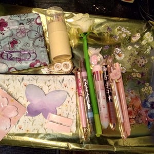 May include: A collection of stationery items including two zippered pouches with floral and butterfly patterns, a cylindrical pencil case, a rose-shaped pen, several multi-colored pens, a butterfly-shaped sticky note, and a cherry blossom sticky note.
