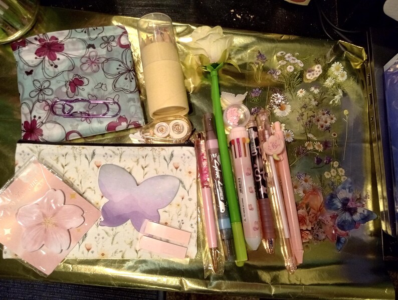 May include: A collection of stationery items including two zippered pouches with floral and butterfly patterns, a cylindrical pencil case, a rose-shaped pen, several multi-colored pens, a butterfly-shaped sticky note, and a cherry blossom sticky note.