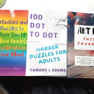 May include: Three paperback books by Tamara L. Adams. The left book has a rainbow cover with the text Activities and Puzzles to Cheer You UP Relieve Anxiety and Brighten Your Mood. The center book is 100 DOT TO DOT HARDER PUZZLES FOR ADULTS. The right book is ART UP THIS JOURNAL.