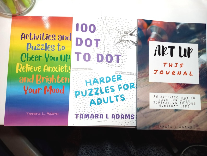 May include: Three paperback books by Tamara L. Adams. The left book has a rainbow cover with the text Activities and Puzzles to Cheer You UP Relieve Anxiety and Brighten Your Mood. The center book is 100 DOT TO DOT HARDER PUZZLES FOR ADULTS. The right book is ART UP THIS JOURNAL.