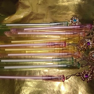 May include: A collection of translucent pens in shades of pink, yellow, and green. Each pen features a white ink cartridge and a gold-toned metal topper shaped like a flower, accented with small embedded crystals in blue, purple, and clear colors.