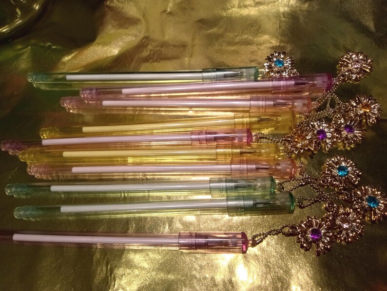 May include: A collection of translucent pens in shades of pink, yellow, and green. Each pen features a white ink cartridge and a gold-toned metal topper shaped like a flower, accented with small embedded crystals in blue, purple, and clear colors.