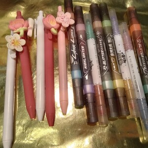 May include: A collection of writing instruments including five pens with floral toppers in shades of white, pink, and red, alongside six markers labeled with text such as Cyber-Lumen, Highlighter, and Glitter, featuring dark and metallic barrel designs.