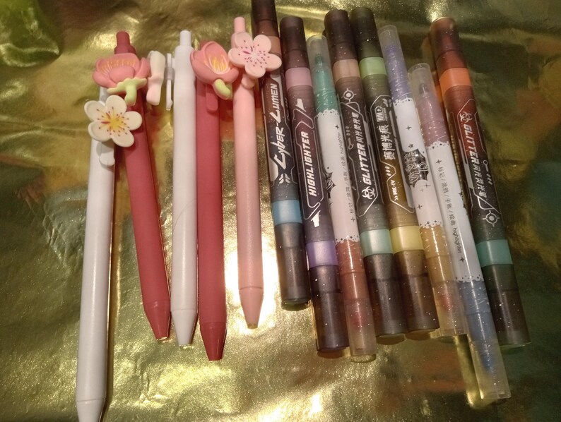 May include: A collection of writing instruments including five pens with floral toppers in shades of white, pink, and red, alongside six markers labeled with text such as Cyber-Lumen, Highlighter, and Glitter, featuring dark and metallic barrel designs.