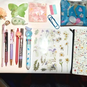 May include: A collection of stationery items including a flower-topped pen, a green butterfly sticky note, cherry blossom sticky notes, a blue paperclip, a blue pouch with a deer print, various pens, and a floral sticker sheet with a matching pencil case.