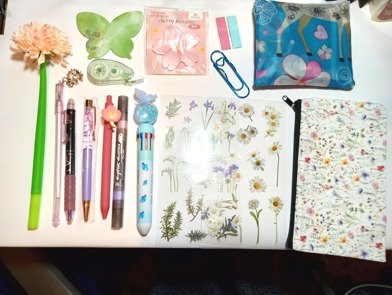 May include: A collection of stationery items including a flower-topped pen, a green butterfly sticky note, cherry blossom sticky notes, a blue paperclip, a blue pouch with a deer print, various pens, and a floral sticker sheet with a matching pencil case.