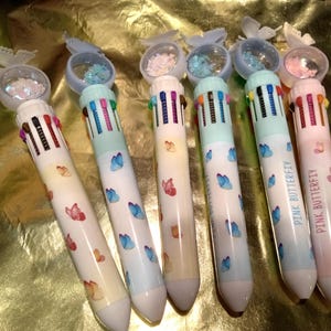 May include: Six multi-color ink pens featuring a butterfly topper with loose glitter inside a clear dome. Each pen has a white barrel decorated with small butterfly illustrations and a row of retractable colored ink switches at the top.