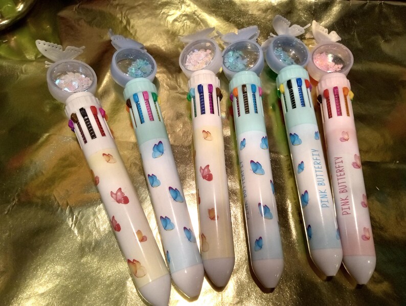 May include: Six multi-color ink pens featuring a butterfly topper with loose glitter inside a clear dome. Each pen has a white barrel decorated with small butterfly illustrations and a row of retractable colored ink switches at the top.
