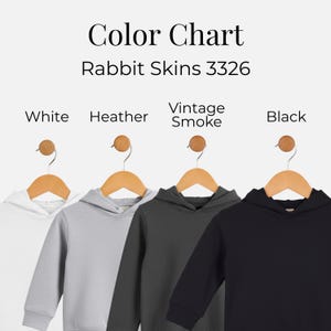 May include: Color chart for Rabbit Skins 3326 hoodies. The image displays four children's hoodies in white, heather gray, vintage smoke gray, and black. Each hoodie hangs on a wooden hanger with a wooden knob.