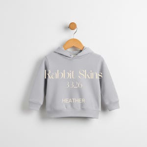 May include: A light gray heather hoodie with a drawstring hood, long sleeves, and ribbed cuffs and hem. The text "Rabbit Skins 3326 HEATHER" is printed on the front. The hoodie is hanging on a wooden hanger.