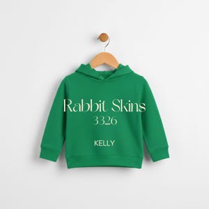 May include: A green hooded sweatshirt with the text "Rabbit Skins 3326 KELLY" in white. The sweatshirt is hanging on a wooden hanger against a white background. The garment has long sleeves and a kangaroo pocket.