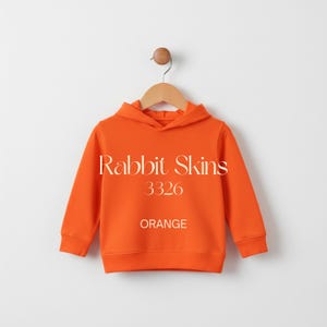 May include: Bright orange hooded sweatshirt with the text "Rabbit Skins 3326 ORANGE". The sweatshirt is hanging on a wooden hanger against a white background. The sweatshirt has long sleeves and a ribbed hem and cuffs.