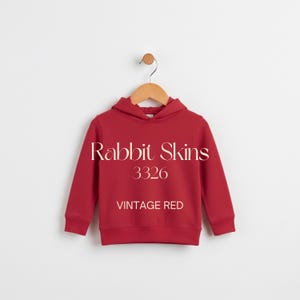 May include: A red hooded sweatshirt with the text "Rabbit Skins 3326 VINTAGE RED" is hanging on a wooden hanger. The sweatshirt is a solid color and has a drawstring hood and ribbed cuffs and hem.