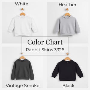 May include: A color chart displays four children's hoodies in white, heather gray, vintage smoke, and black. The hoodies are hanging on wooden hangers. The text "Color Chart" and "Rabbit Skins 3326" are in the center.